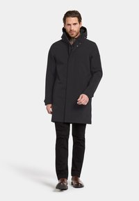 Svart vattentät jacka med huva, med dragkedja, sidofickor och rak silhuett, kombinerad med svarta byxor och bruna skor.