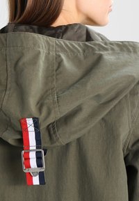 Olivově zelená bunda s kapucí, která má kontrastní pruhovaný textilní popruh s kovovou sponou na zadní straně, hladkou texturu a odolný materiál.