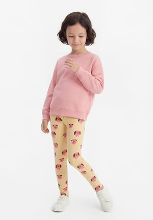 Παιδί που φοράει ροζ φούτερ, μπεζ κολάν με ροζ πρόσωπα σκύλων και λευκά αθλητικά παπούτσια, στέκεται μπροστά σε λευκό φόντο.