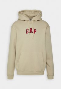 Sudadera con capucha beige hecha de algodón, con logo "GAP" bordado en rojo en el pecho, bolsillo frontal tipo canguro y capucha con cordón ajustable.