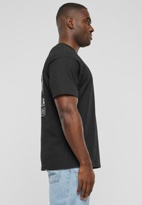 Sort bomulds t-shirt vist på en person, med korte ærmer og rund halsudskæring, med et grafisk print på ryggen.