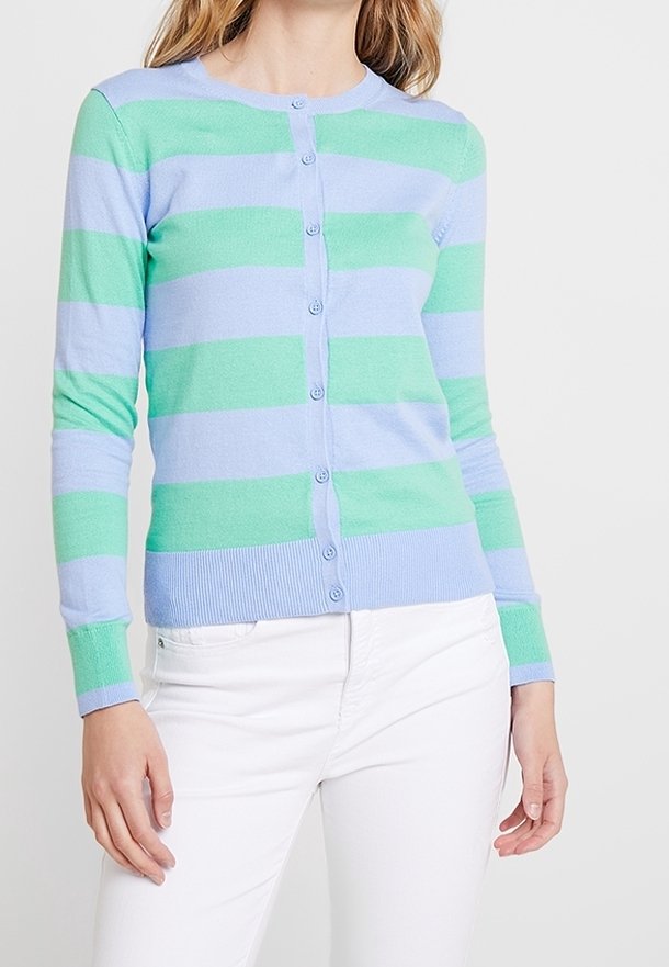 Femme portant un cardigan boutonné à rayures horizontales bleu pastel et vert avec manches longues et pantalon blanc.