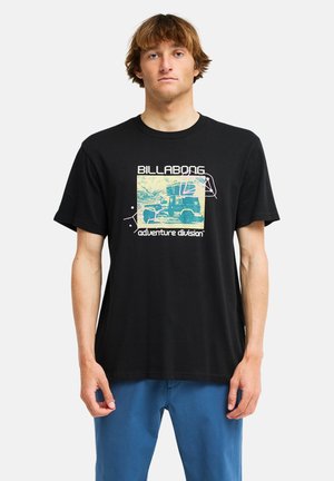 Junger Mann mit hellbraunen Haaren, der ein schwarzes Billabong-T-Shirt mit einem Vintage-Fahrzeug-Motiv und blauen Hosen trägt, steht vor einem weißen Hintergrund.