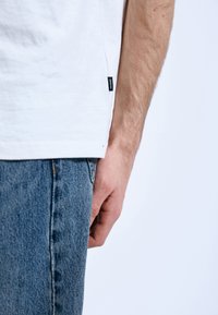 Bílý bavlněný tričko s postranními štítky, v kombinaci s modrými džíny, zobrazuje detail ruky a dolní části oděvu.