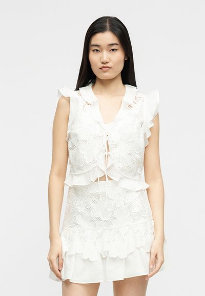 Νέα γυναίκα με μακριά σκούρα μαλλιά φοράει αμάνικο λευκό φόρεμα δαντέλα με βολάν, στέκεται σε απλό φωτεινό φόντο.