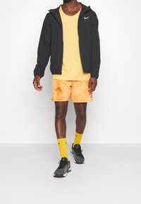 Man wearing black zip-up jacket, yellow t-shirt, orange tie-dye shorts, yellow socks, and black athletic shoes, standing against a white background.