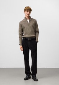 Sweter z dzianiny w brązowym, wielopatternowym wzorze, z kołnierzem na ćwierć zamka. Czarna zamszowa para spodni, dopełniona czarnym skórzanym paskiem i butami.