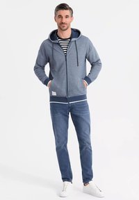 Haut à capuche zippé bleu clair avec une doublure marine, doté de cordons de serrage, associé à une chemise rayée et un jean bleu. Porté avec des baskets blanches.