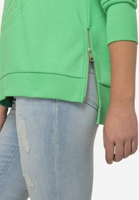 Sudadera verde con cremallera lateral, puños y dobladillo acanalados, combinada con jeans descoloridos de color azul claro, que presentan detalles de rodillas deshilachadas.