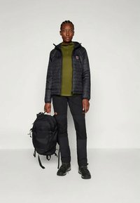 Musta eristävä takki oliivi vihreän pitkähköhihaisen paidan päällä, mustat housut jossa reisi- ja polvitaskut, sekä mustat vaelluskengät. Musta reppu vieressä.