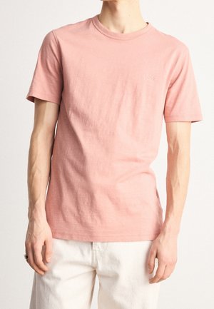 Mand iført en ensfarvet lyserød kortærmet T-shirt og hvide bukser mod en ensfarvet lys baggrund.