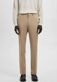 Pantalones beige a medida con corte recto, frente plano, trabillas para cinturón, bolsillos laterales y textura sutil, combinados con zapatos negros y un suéter ligero.