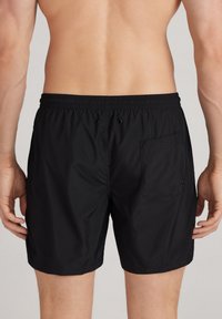 Pantalones de baño negros con cintura elástica, cordón de ajuste y un bolsillo de parche en el lado derecho. Tejido suave con una textura ligera.