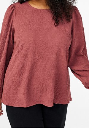 Vrouw die een gestructureerde stofkleurige roze blouse met lange mouwen en pofschouders draagt, en zwarte broek, getoond van nek tot heupen.