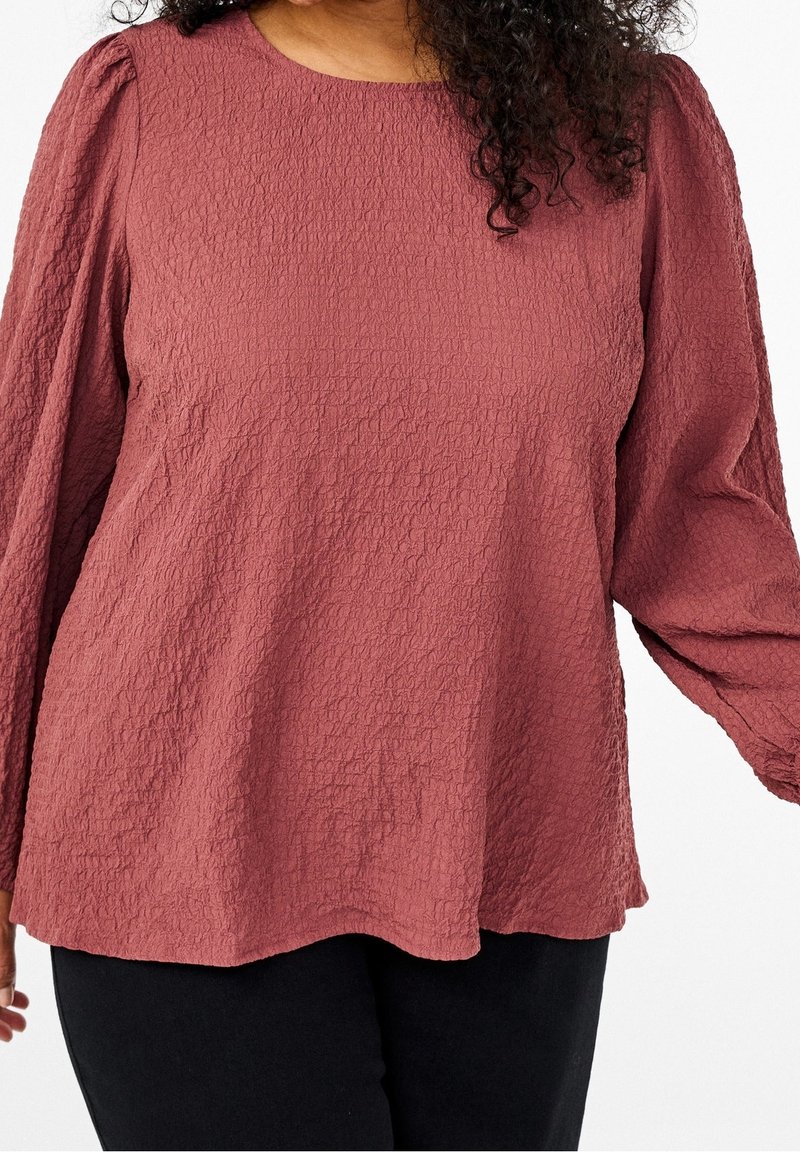 Femme portant un chemisier texturé rose poudré à manches longues avec épaules bouffantes et pantalon noir, vue du cou aux hanches.