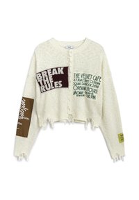 Pull en blanc, raccourci, avec des bords effilochés, présentant des patchs de tissu variés en marron et vert avec des détails textuels, sur un fond en maille texturée.