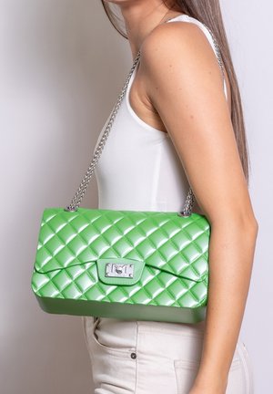 Femme portant un haut sans manches blanc et un pantalon clair, portant un sac à main matelassé vert brillant avec une bandoulière en chaîne argentée sur l'épaule.