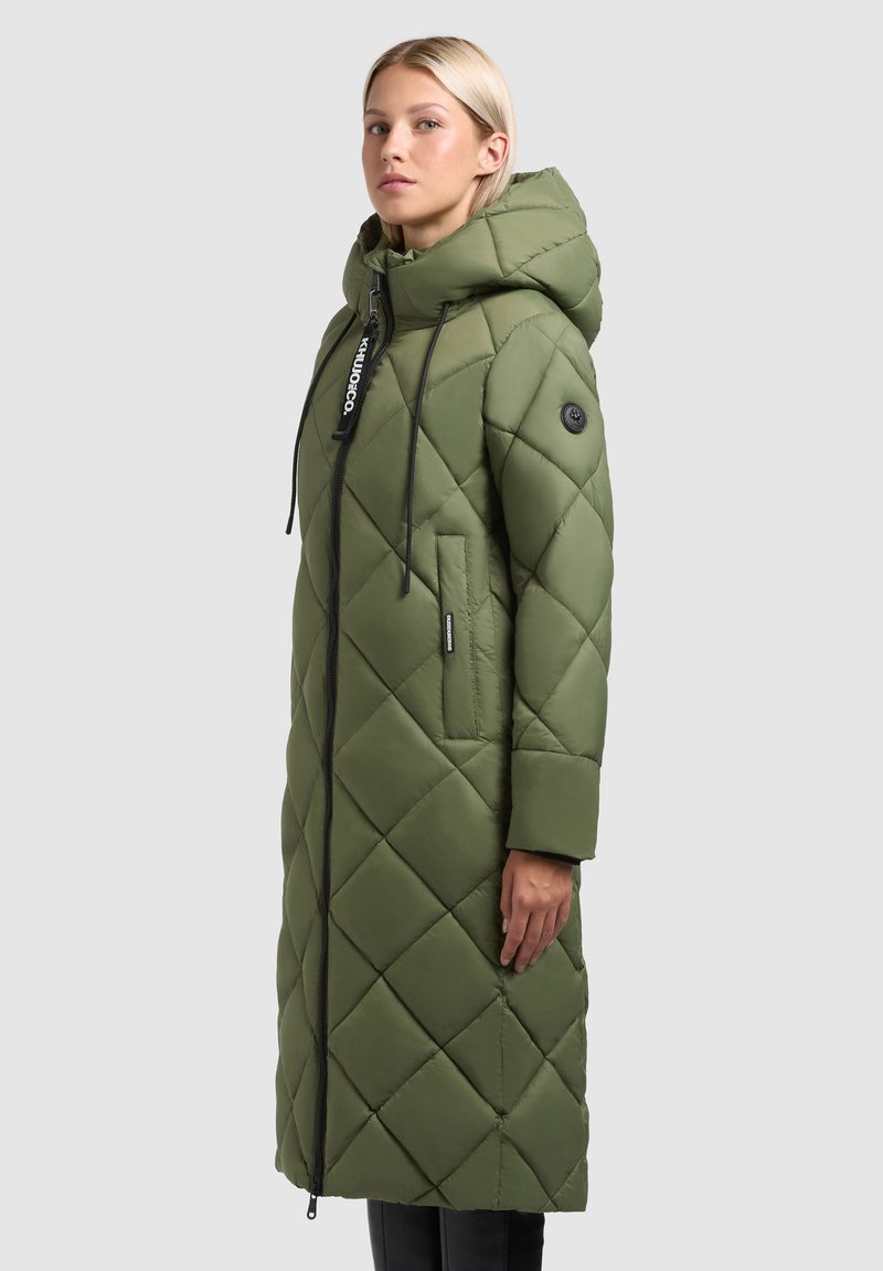 Palton lung, verde măsliniu, căptușit, cu glugă, având un fermoar frontal, accente negre și buzunare laterale. Model texturat în formă de romb pe întreaga suprafață.