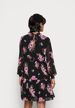 Woman with short dark hair wearing a black long-sleeve dress with pink floral patterns, shown from the back against a plain background.