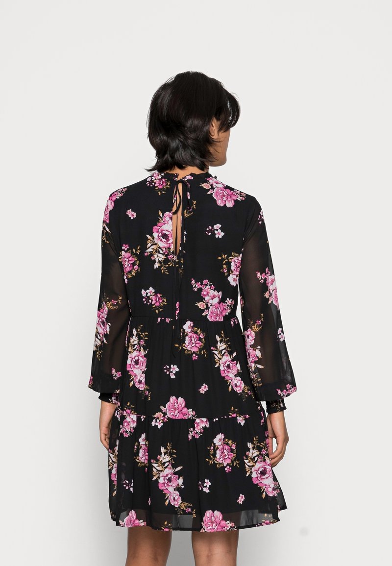 Femme aux cheveux courts et foncés portant une robe noire à manches longues avec des motifs floraux roses, vue de dos sur un fond uni.