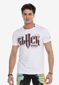 Weißes Baumwoll-T-Shirt mit rundem Hals, das auf der Vorderseite mit kräftigem schwarzem und rotem grafischen Text bedruckt ist. Kurze Ärmel, kombiniert mit bunten Batik-Hosen.