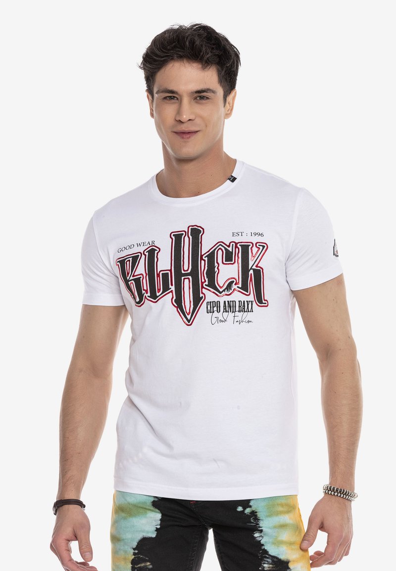 Weißes Baumwoll-T-Shirt mit rundem Hals, das auf der Vorderseite mit kräftigem schwarzem und rotem grafischen Text bedruckt ist. Kurze Ärmel, kombiniert mit bunten Batik-Hosen.