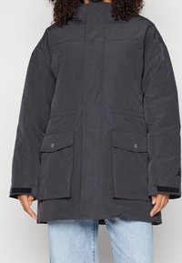 Mörkgrå parka-jacka gjord av ett strukturerat tyg, med hög krage, två stora framfickor och svarta detaljer vid ärmsluten.