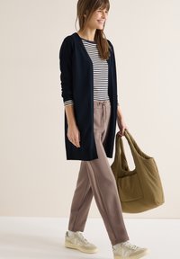 Femme portant une chemise rayée, un cardigan bleu marine, un pantalon mauve, des baskets blanches, et portant un sac cabas vert olive, marchant et souriant.