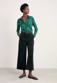 Žalias raštuotas marškinėliai su ilgomis rankovėmis ir apykakle, derinami su tamsiai žaliais aksominio plačių kelnių ir tamsiais plokščiais batais.