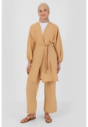 Beżowy płaszcz w stylu kimono z długimi, balonowymi rękawami, z pasującym paskiem w talii, zestawiony z luźnymi beżowymi spodniami i brązowymi sandałami.