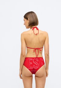 Κόκκινο μπικίνι σετ με λαιμόκοψη τύπου halter και ρυθμιζόμενες κρέμες, καθώς και ψηλόμεσο κάτω μέρος που παρουσιάζει το γυαλιστερό λογότυπο "DIESEL" σε αντίθετη υφή.