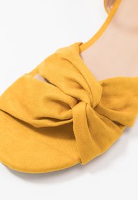 Keltainen mokkanahkakengät, joissa on solmittu muotoilu päällä. Littee pohja ja avoin varvas, jossa on sileä pinta ja vähäinen varustus.