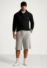 Άντρας που φοράει μαύρη κουκούλα, μαύρο καπέλο, γκρι σορτς, μπεζ καλτσόν και λευκά αθλητικά παπούτσια, στέκεται με το ένα χέρι στην τσέπη σε απλό φόντο.