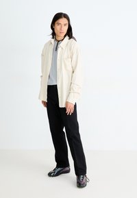 Cămiză bej deschis, în dungi, peste un top gri; pantaloni negri; adidași multicolorați cu talpă texturată; model în picioare.