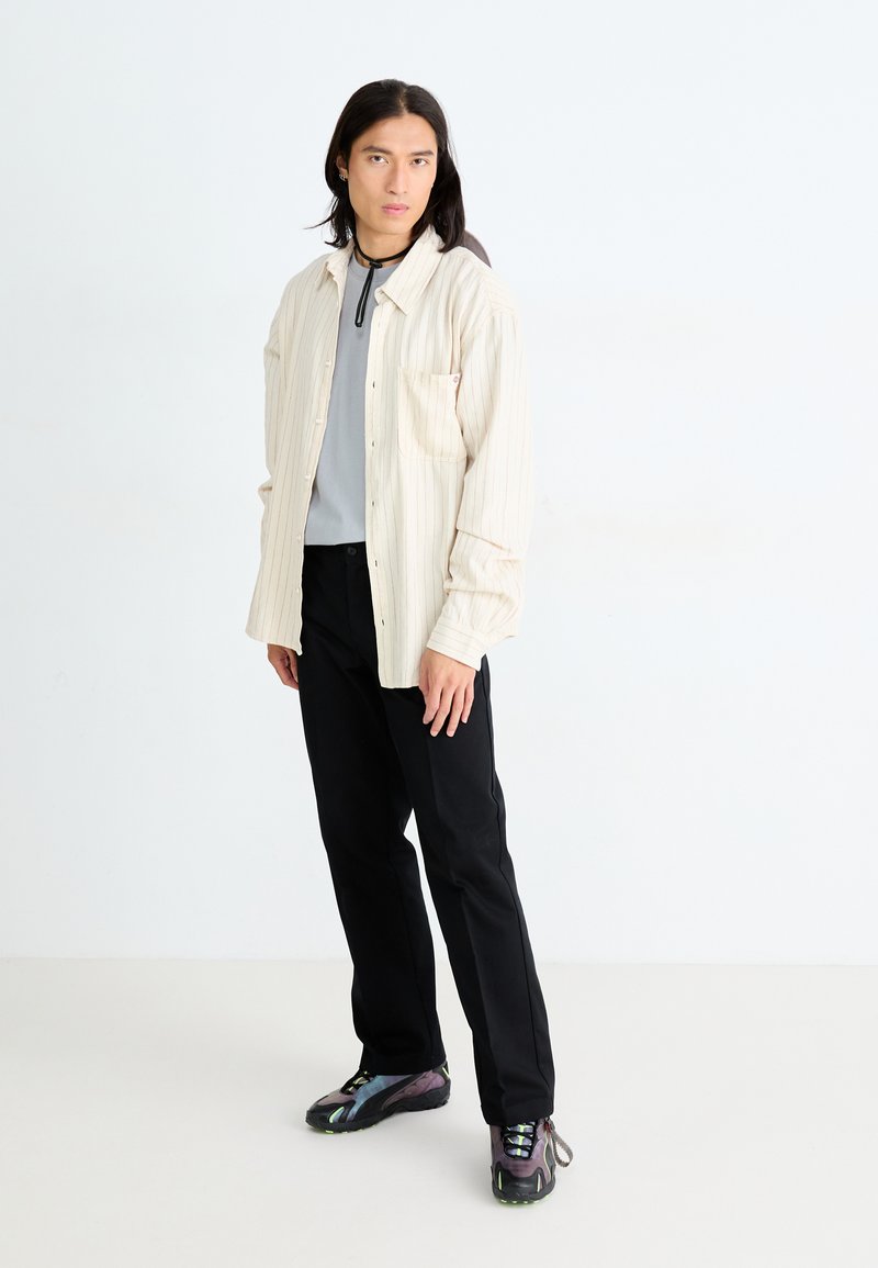 Helles beige, gestreiftes Hemd über einem grauen Oberteil; schwarze Hose; mehrfarbige Sneaker mit strukturiertem Sohlen; stehendes Model.