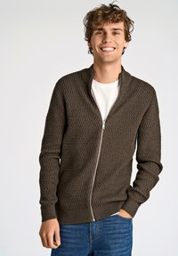 Cardigan zip-up marrone con trama, colletto alto e motivo geometrico. Indossato sopra una maglietta bianca, abbinato a jeans blu.