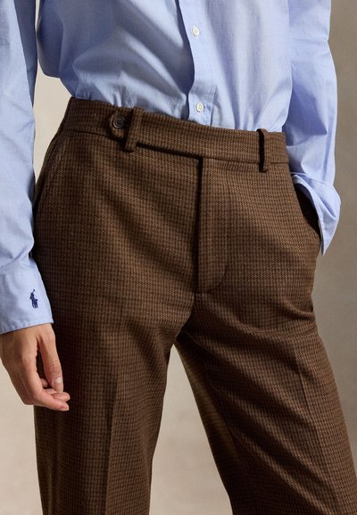 Homme portant un pantalon marron texturé et une chemise bleu clair boutonnée avec un petit logo brodé sur le poignet.