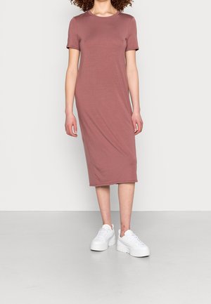 Kvinde iført en støvet rosa, kortærmet, midi lang t-shirt kjole med hvide platåsneakers, stående mod en ensfarvet lys baggrund.