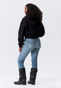 Γυναίκα με σγουρά μαλλιά που φοράει μαύρο φούτερ με κουκούλα, ανοιχτό μπλε τζιν και μαύρες μπότες μέχρι το μέσο της γάμπας, στέκεται ελαφρώς πλαγιασμένη σε ένα απλό φόντο.