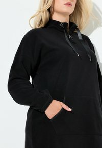 Svart fleece-pullov med hög krage, zippered nacke, justerbara snoddar och framficka. Mjuk textur och avslappnad passform.