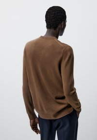 Homme aux cheveux bruns bouclés, portant un pull marron à manches longues et un pantalon foncé, debout de dos sur un fond uni.