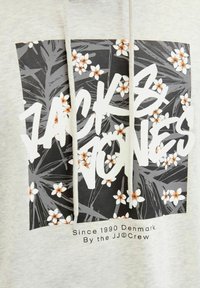 Svijetlosivi dukserica s kapuljačom s crnim cvjetnim grafikom koja sadrži bijele i narančaste cvjetove, a ispod je centriran podebljani natpis "ZACK & JONES".