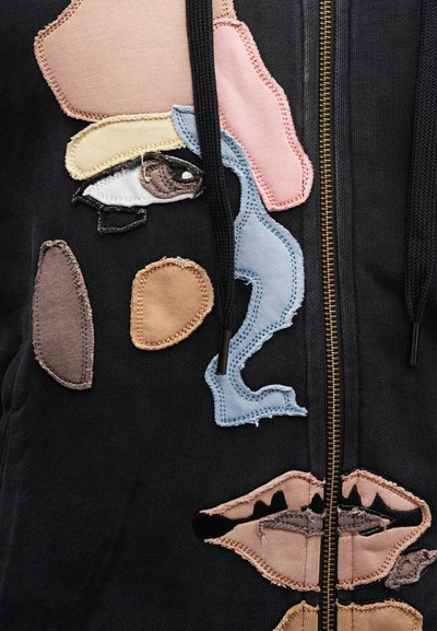 Μαύρο φούτερ με φερμουάρ, διακοσμημένο με διάφορα υφασμάτινα μπαλώματα σε ακανόν ιστες μορφές και χρώματα, περιλαμβάνοντας μάτια και χείλη, με αντίθετες ραφές.