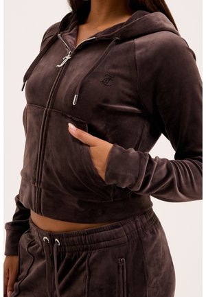 Femme portant un sweat à capuche zippé en velours marron foncé et un pantalon à cordon assorti, la main dans la poche du sweat.