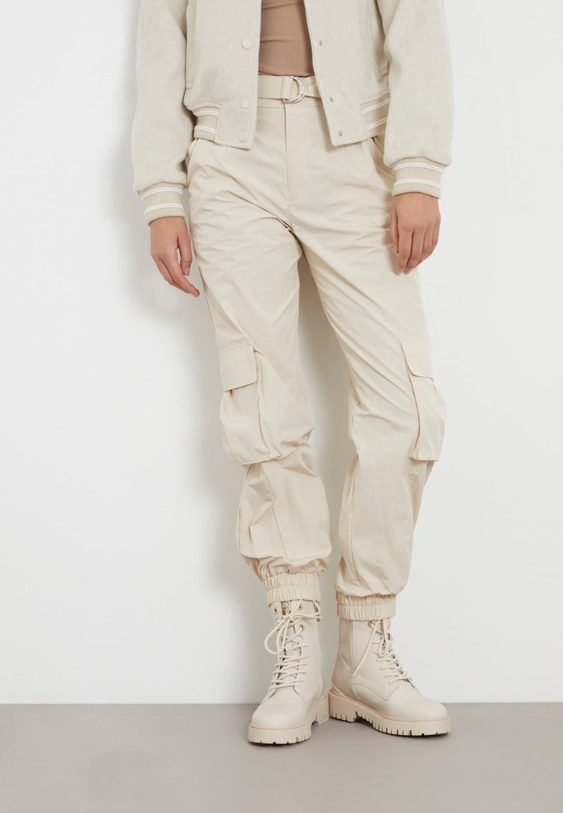 Personne portant un pantalon cargo beige avec des poignets élastiques, une ceinture, des bottines à lacets assorties et une veste beige par-dessus un haut couleur sable.
