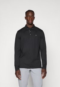 Μαύρο μακρυμάνικο polo μπλουζάκι με γιακά, τρία κουμπιά και ελάχιστο λογότυπο. Φτιαγμένο από μαλακό ύφασμα, σε συνδυασμό με ανοιχτό γκρι παντελόνι.