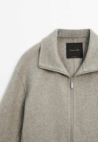 Szara kurtka z mieszanki wełny z zamkiem błyskawicznym, z kołnierzem do złożenia i srebrnym zamkiem. Oznaczona czarną metką z białym tekstem.