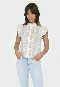 Blouse en dentelle blanche avec des manches courtes à volants, présentant des motifs verticaux et une fermeture à boutons à l'avant. Associée à un jean bleu clair.
