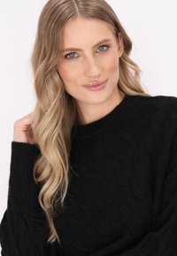 Schwarzer Strickpullover mit einem strukturierten Rautenmuster und rundem Halsauschnitt, getragen von einem Model mit welligem Haar und sanftem Make-up.