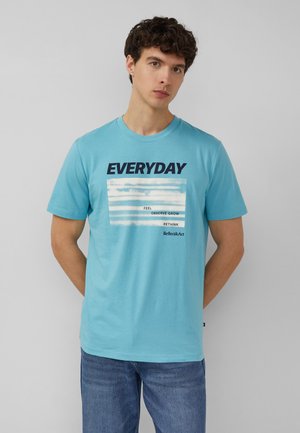 Mężczyzna w jasnoniebieskim T-shircie z napisem „EVERYDAY” i mniejszym tekstem na białych paskach, w niebieskich dżinsach, stoi przy szarej ścianie.